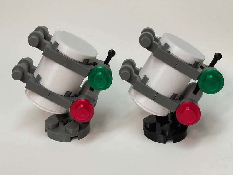 May include: Two miniature, futuristic-looking models. Each has a white cylindrical body, gray supports, and a base. One has a red and green light, the other has a red and green light. The bases are gray and black.