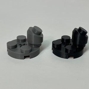 May include: Two 3D-printed plastic objects, one gray and one black, resembling stylized figures. Each has a circular base with three raised circular elements and a central, angled structure with a cylindrical top. The objects are on a white surface.