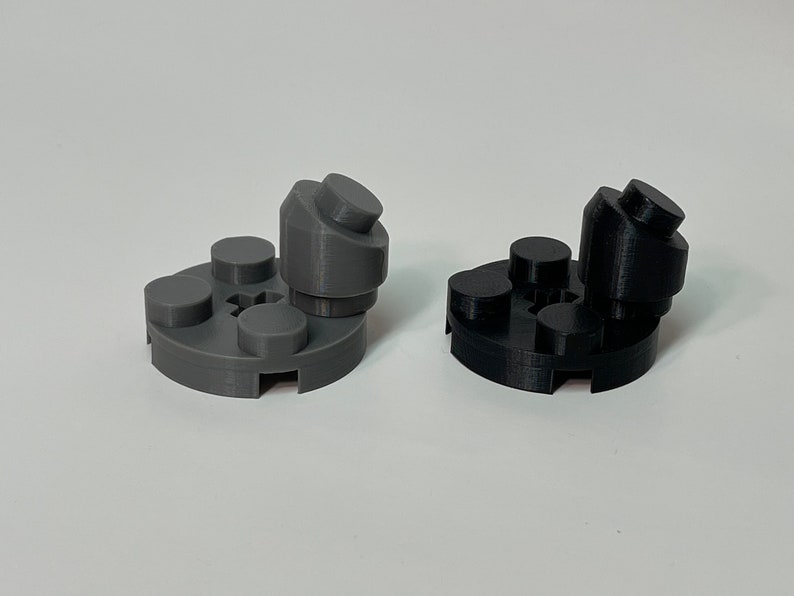 May include: Two 3D-printed plastic objects, one gray and one black, resembling stylized figures. Each has a circular base with three raised circular elements and a central, angled structure with a cylindrical top. The objects are on a white surface.