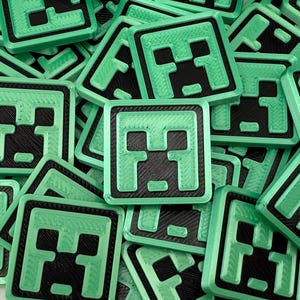 May include: A collection of square, green and black Minecraft-themed badges. Each badge features a pixelated face design with a green border. The badges are scattered across a surface, creating a textured pattern.