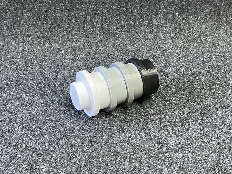 May include: A multi-colored bottle stopper with a stepped design. The stopper is composed of white, light gray, dark gray, and black sections. The object is cylindrical and appears to be made of plastic.