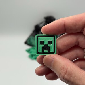 May include: A small, square, green and black Minecraft Creeper face emblem. The emblem is held between fingers against a white background. The Creeper face is a pixelated design with a green border.