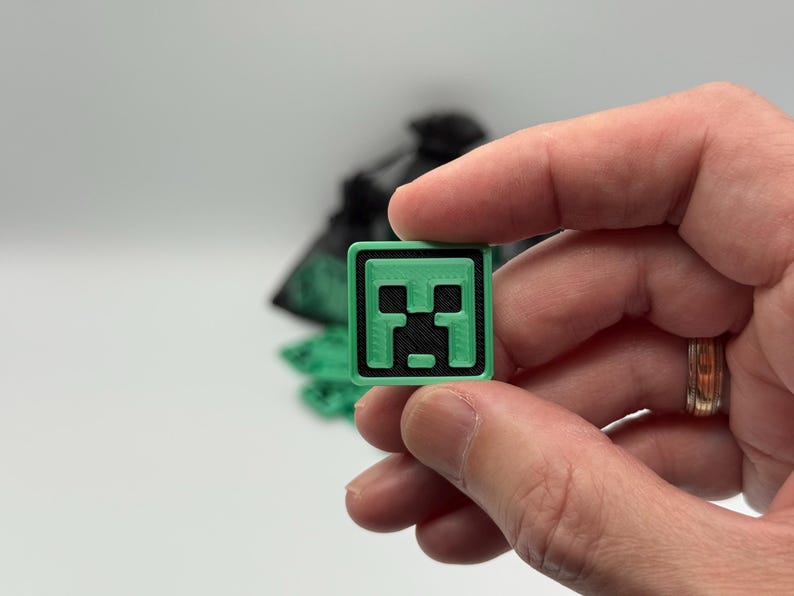 May include: A small, square, green and black Minecraft Creeper face emblem. The emblem is held between fingers against a white background. The Creeper face is a pixelated design with a green border.