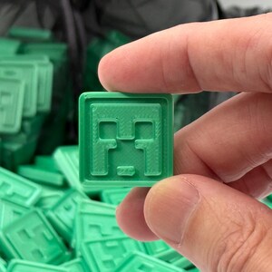Puede incluir: Una baldosa cuadrada verde con un dise&ntilde;o de cara pixelada, sostenida entre los dedos. La baldosa forma parte de una colecci&oacute;n m&aacute;s grande de baldosas verdes similares, lo que sugiere un tema de juego o artesan&iacute;a. El dise&ntilde;o recuerda a un personaje popular de videojuegos.