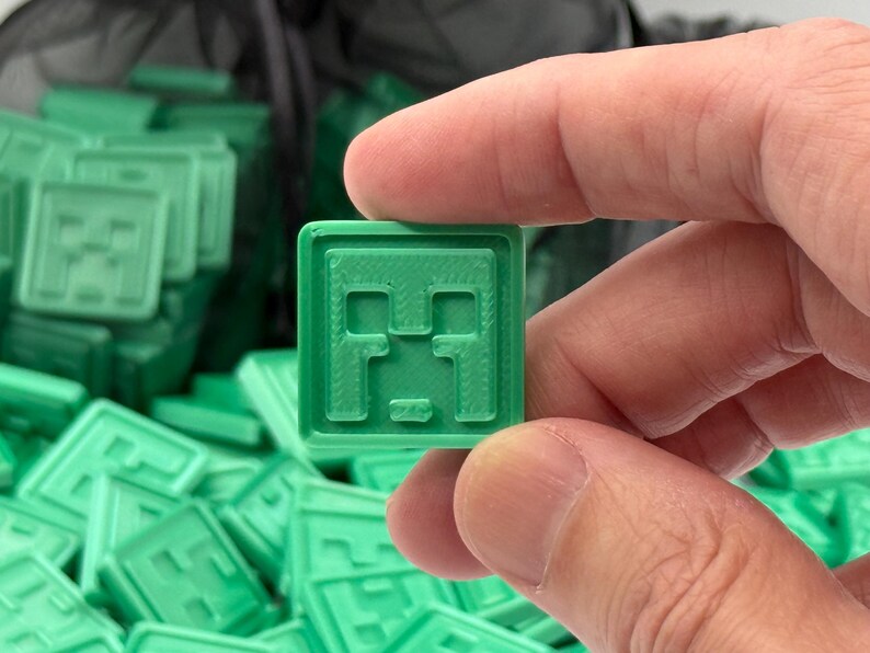 Puede incluir: Una baldosa cuadrada verde con un dise&ntilde;o de cara pixelada, sostenida entre los dedos. La baldosa forma parte de una colecci&oacute;n m&aacute;s grande de baldosas verdes similares, lo que sugiere un tema de juego o artesan&iacute;a. El dise&ntilde;o recuerda a un personaje popular de videojuegos.