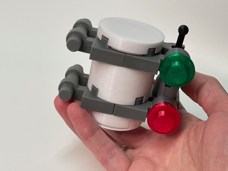 May include: A 3D-printed device with a white cylindrical body, gray frame, and a white top. It features a green and a red light, and a black antenna-like element. The device is held in a hand.
