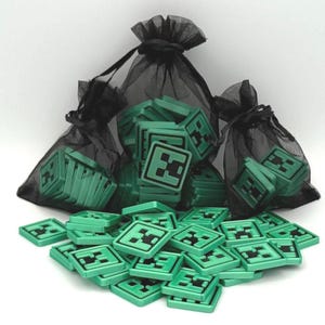 May include: Green square game pieces with a black Creeper face design are scattered on a white surface. Some pieces are in black mesh bags. The pieces are likely for a board game or collectible.
