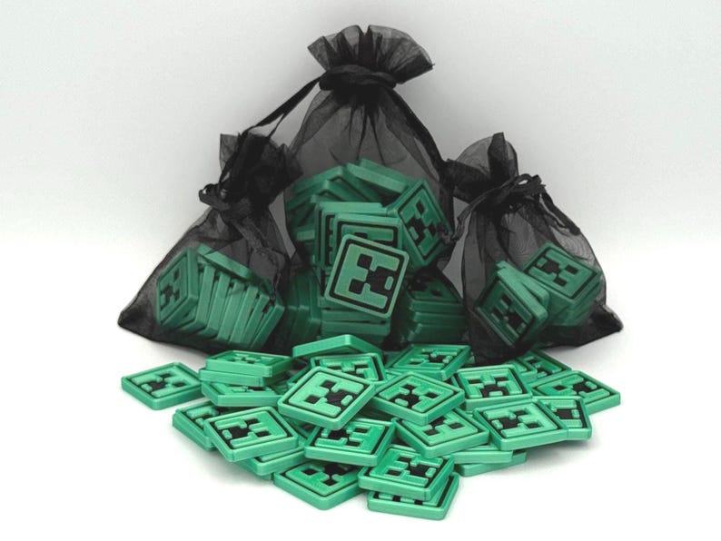 May include: Green square game pieces with a black Creeper face design are scattered on a white surface. Some pieces are in black mesh bags. The pieces are likely for a board game or collectible.