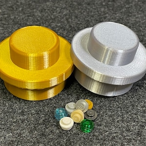 May include: Two large, 3D-printed Lego-style bricks, one gold and one silver. A collection of smaller Lego pieces in various colours, including clear blue, white, and green, are scattered below the larger bricks. The items are on a grey surface.