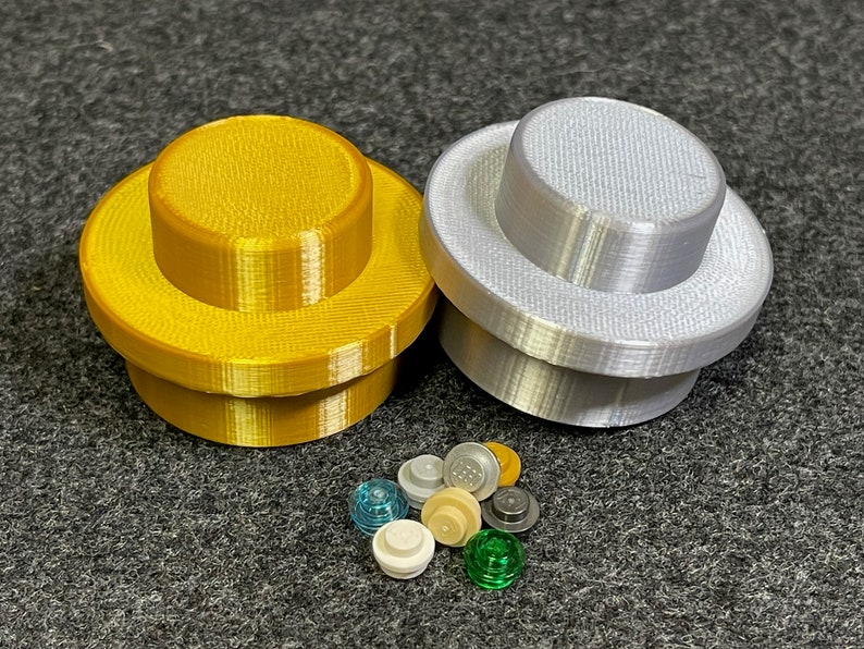 May include: Two large, 3D-printed Lego-style bricks, one gold and one silver. A collection of smaller Lego pieces in various colours, including clear blue, white, and green, are scattered below the larger bricks. The items are on a grey surface.