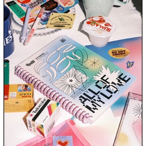 May include: A pink and blue spiral bound notebook with the text "Roilli Notebook" and "All of My Love" in black lettering. The notebook is open and lying on a table with other items, including a camera, film, and a small container.