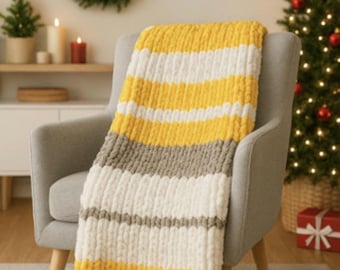 May include: A chunky knit blanket draped over a gray armchair. The blanket features horizontal stripes of yellow, white, and gray. The scene is set in a cozy living room with a Christmas tree and wreath.