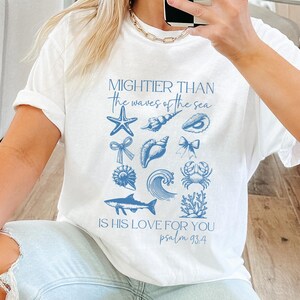 May include: White t-shirt with blue marine-themed graphics and text that reads "Mightier than the waves of the sea, is his love for you, psalm 93:4." The design includes starfish, shells, and a wave.