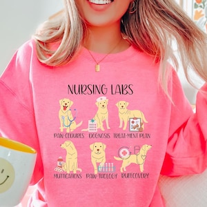 Può includere: Felpa rosa con una grafica di un cane Labrador Retriever in diverse pose con il testo "Nursing Labs" e le parole "Paw-cedures, Dognosis, Treat-ment Plan, Muttications, Paw-thology, Ruffcovery" sotto.