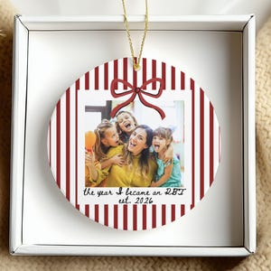 May include: A round ceramic ornament featuring a red and white vertical striped border, a red bow graphic at the top, and a central photograph of four people. The text reads: the year I became an RBT est. 2026. It hangs by a gold string.