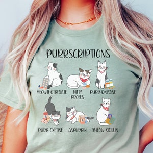 以下が含まれることがあります： 猫のグラフィックと「PURRSCRIPTIONS」というテキストが書かれた薄い緑色のTシャツ。下に猫をテーマにした薬のリストがあります。
