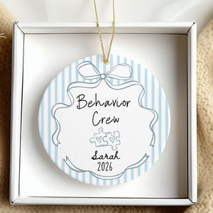 May include: A round white ceramic ornament featuring light blue vertical stripes, a hand-drawn ribbon border, and the text "Behavior Crew" above two puzzle pieces with hearts. The name "Sarah" and the year "2026" are printed below. Gold string attached.