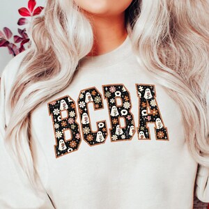 Könnte beinhalten: Ein sandfarbenes Sweatshirt mit Rundhalsausschnitt und den Buchstaben BCBA in einer fetten Blockschrift. Die Buchstaben sind mit einem schwarzen Muster aus kleinen weißen Geistern, Blumen und Sternen gefüllt und braun umrandet.