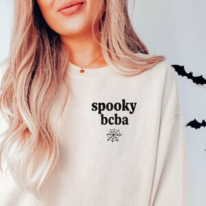 Könnte beinhalten: Ein sandfarbenes Sweatshirt mit Rundhalsausschnitt und schwarzem Schriftzug "spooky bcba" auf der linken Brustseite, unter dem sich eine kleine schwarze Spinnennetz-Grafik befindet. Das Kleidungsstück hat lange Ärmel sowie gerippte Bündchen.
