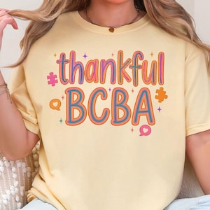 Könnte beinhalten: Ein hellgelbes Kurzarm-T-Shirt mit dem Schriftzug thankful BCBA in einer mehrfarbigen Retro-Schriftart. Das Design enthält kleine Puzzleteil-Symbole, Sprechblasen und Sternformen in Rosa-, Orange- und Blautönen auf der Vorderseite.