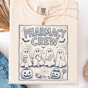 K&ouml;nnte beinhalten: Ein elfenbeinfarbenes T-Shirt mit einem dunkelblauen Grafikdruck und dem Text PHARMACY CREW. Das Design zeigt vier Geister mit Apothekenartikeln, einen K&uuml;rbis, einen S&uuml;&szlig;igkeiten-Eimer, Spinnweben, Flederm&auml;use und verschiedene Pillen im gesamten Motiv.