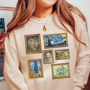 Puede incluir: Una sudadera beige con imágenes enmarcadas de pinturas de Vincent van Gogh. La obra de arte incluye "La noche estrellada", retratos y naturalezas muertas florales. La sudadera tiene cuello redondo y mangas largas.