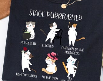 May include: Navy blue t-shirt with the text "STAGE PURRFORMER" and illustrations of cats in theatrical roles. The shirt features cats in roles such as "Meowcbeth" and "Romeow & Juliet". A gold necklace is draped on the shirt.
