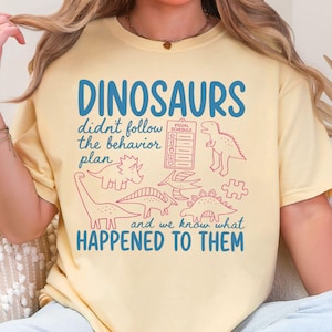 May include: A pale yellow t-shirt featuring blue text that reads DINOSAURS didn't follow the behavior plan and we know what HAPPENED TO THEM. The design includes red line art of various dinosaurs, a visual schedule chart, and a puzzle piece.