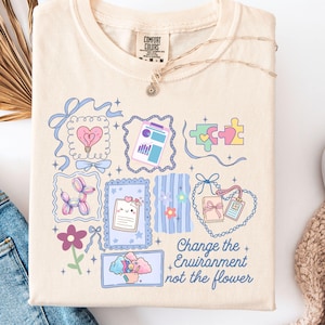 Puede incluir: Camiseta Comfort Colors color marfil con un diseño gráfico colorido. El diseño incluye varias ilustraciones como un corazón, piezas de rompecabezas y un animal de globo. El texto en la camiseta dice "Change the Environment not the flower."