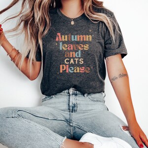 K&ouml;nnte beinhalten: Ein dunkelgraues T-Shirt mit dem Text "Autumn leaves and cats please" in bunten Buchstaben.