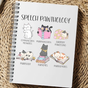 May include: A spiral-bound notebook with the title "SPEECH PAWTHOLOGY" and illustrations of cats. The notebook features various cat-themed puns related to speech therapy, such as "Communication Meowdels" and "Purrnunciation". The notebook is on a woven surface.