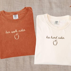 May include: Two crewneck t-shirts. The yam-coloured shirt features the text "her apple cider" with an apple graphic. The ivory-coloured shirt features the text "his hard cider" with an apple graphic. Both shirts have a Comfort Colors brand label.