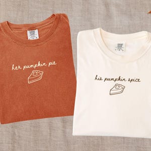 May include: Two crew neck t-shirts. The left shirt is yam-coloured with white text reading 'her pumpkin pie' above a slice of pie graphic. The right shirt is ivory-coloured with brown text reading 'his pumpkin spice' above a slice of pie graphic.