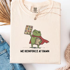 May include: An ivory t-shirt featuring a graphic of a green frog wearing a red cape, holding a grid board with colored circles and a clipboard. Below the frog, the text reads WE REINFORCE AT DAWN. The shirt has a Comfort Colors brand label at the neck.