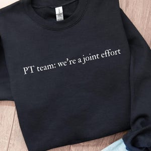 May include: A black long-sleeve crewneck sweatshirt featuring the white printed text "PT team: we're a joint effort" across the chest. The garment has a ribbed neckline, cuffs, and hem, with a standard fit and a visible brand label at the neck.