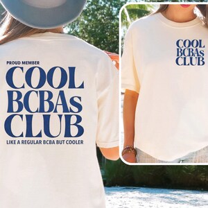 Könnte beinhalten: Ein elfenbeinfarbenes Kurzarm-T-Shirt mit dunkelblauem Text. Auf der Rückseite stehen die Worte PROUD MEMBER COOL BCBAs CLUB LIKE A REGULAR BCBA BUT COOLER. Auf der linken Brustseite befindet sich der kleinere Text COOL BCBAs CLUB in Dunkelblau.