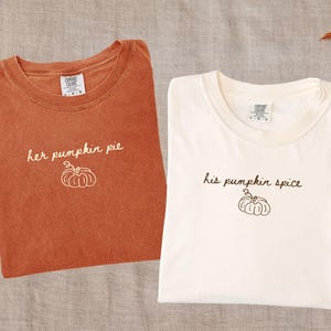 May include: Two crew neck t-shirts. The left shirt is yam-coloured with white text reading 'her pumpkin pie' and a white pumpkin outline. The right shirt is ivory with brown text reading 'his pumpkin spice' and a brown pumpkin outline.
