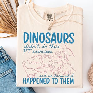 May include: An ivory short-sleeve t-shirt featuring blue text that reads "DINOSAURS didn't do their PT exercises and we know what HAPPENED TO THEM" alongside pink line-art illustrations of various dinosaurs, a dumbbell, and an exercise ball.