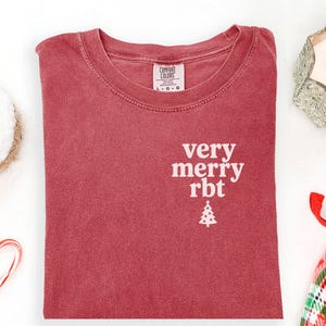 May include: A crimson red short-sleeve t-shirt with a crew neckline. The chest features white text that reads very merry rbt above a small white Christmas tree graphic. The interior neck label displays the brand name Comfort Colors and size L-G.