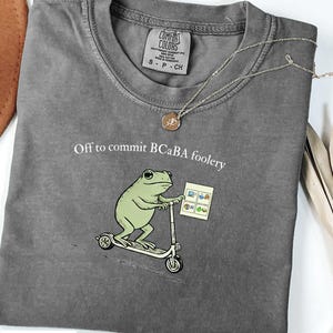 May include: A gray Comfort Colors t-shirt with the text "Off to commit BCABa foolery" and a cartoon frog riding a scooter. The frog is holding a paper with images. A gold necklace is draped across the shirt.