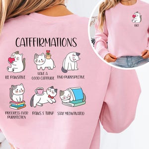 Op de afbeelding: Lichtroze sweatshirt met de tekst "CATFFIRMATIONS" en cartoonkattenillustraties. De illustraties tonen katten met harten, planten, boeken en koffiekopjes. Er staat ook een kleine kattenafbeelding op de linkerbovenborst.