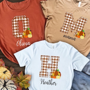 Könnte beinhalten: Drei weiße T-Shirts mit braunen und weißen karierten Buchstaben, Kürbissen und Namen. Die Shirts sagen "Olivia", "mama" und "Heather".