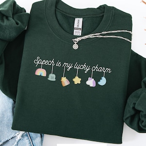 Op de afbeelding: Bostgroen sweatshirt met de tekst "Speech is my lucky charm" in witte letters. Onder de tekst staan kleurrijke bedels, waaronder een regenboog, een bel en een ster. Een zilveren ketting met een zonnehanger ligt op het sweatshirt.