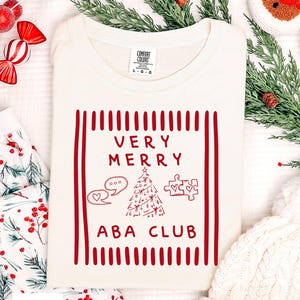 May include: An ivory t-shirt featuring a red graphic print with the text VERY MERRY ABA CLUB. The design includes a Christmas tree, speech bubbles with a heart, and puzzle pieces, all framed by vertical red stripes on the left and right sides.