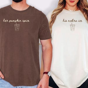 May include: A dark brown t-shirt with the text her pumpkin spice and a line art iced coffee graphic, alongside an ivory t-shirt with the text his extra ice and a line art iced coffee graphic. Both shirts feature short sleeves and crew necklines.