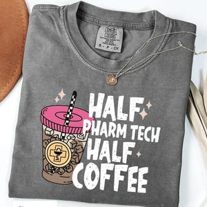 May include: A charcoal gray crew neck t-shirt featuring a graphic of an iced coffee in a plastic cup with a pink lid and a black and white straw. The text reads HALF PHARM TECH HALF COFFEE. The cup includes a gold circular pharmacy symbol icon.