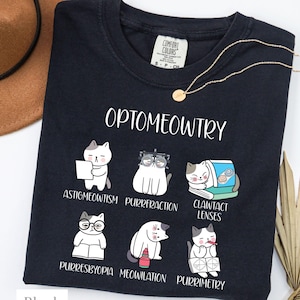 Puede incluir: Camiseta negra con la palabra "Optomeowtry" e ilustraciones de gatos que representan diferentes afecciones oculares. La camiseta también presenta las palabras "Astigmeowtism", "Purrrefraction", "Clawtact Lenses", "Purresbyopia", "Meowilation" y "Purrimetry".
