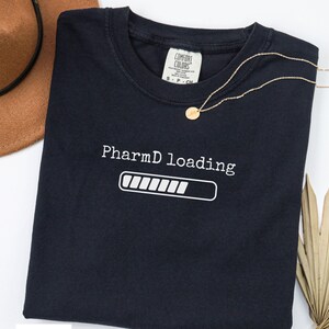 May include: Black t-shirt with the text "PharmD loading" and a loading bar graphic in white. A gold necklace with a small pendant rests on the shirt. A brown hat is visible in the background.