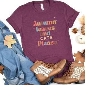 K&ouml;nnte beinhalten: Ein maroonfarbenes T-Shirt mit dem Text "Autumn leaves and cats please" in bunten Buchstaben. Das Shirt wird mit einer blauen Jeans, braunen Stiefeln und Herbstlaub kombiniert.