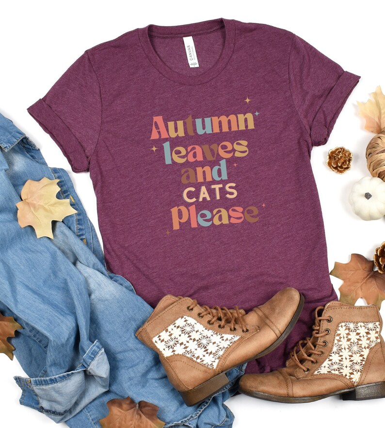K&ouml;nnte beinhalten: Ein maroonfarbenes T-Shirt mit dem Text "Autumn leaves and cats please" in bunten Buchstaben. Das Shirt wird mit einer blauen Jeans, braunen Stiefeln und Herbstlaub kombiniert.